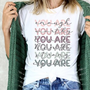 May include: White t-shirt with the text "You are enough, You are worthy, You are loved, You are kind, You are strong, You are capable" printed in a layered, colorful font.