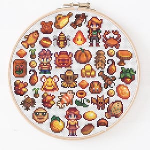 May include: A circular wooden embroidery hoop containing a cross-stitch pattern featuring various pixel art icons, including characters, animals, plants, and food items in shades of orange, brown, yellow, and green on a white fabric background.