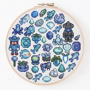 May include: A circular wooden embroidery hoop containing white fabric with a cross-stitch pattern of various blue-toned pixel art icons, including fish, crystals, plants, a small figure in a hat, and a dark-clad character with a wide-brimmed hat.