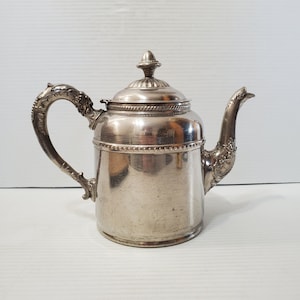 Op de afbeelding: Zilverkleurige theepot met een decoratieve handgreep en tuit. De theepot heeft een deksel met een kleine knop en een gestructureerde band in het midden. De handgreep en tuit hebben sierlijke details, wat wijst op een vintage of antieke stijl.