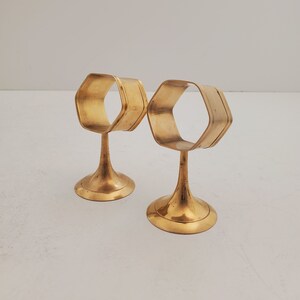 May include: Two gold metal napkin rings with a hexagonal shape and a flared base.