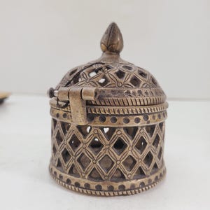 May include: A small, ornate brass box with a hinged lid. The box is decorated with a geometric pattern and has a pointed finial on top.