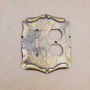 Puede incluir: Placa de cubierta de interruptor de luz y enchufe de estilo antiguo y ornamentado. La placa está hecha de metal texturizado de color dorado con un borde decorativo festoneado. Cuenta con una abertura para un interruptor y dos aberturas para enchufes.