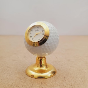 May include: A white golf ball desk clock featuring a gold-toned bezel and a gold-toned pedestal base. The clock face is white with black numerals, gold hands, and the text Goldhill printed in black. The golf ball has a dimpled texture.