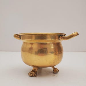 May include: A small, gold-coloured brass pot with two handles and three feet. The pot is round and has a smooth surface.