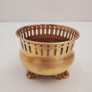 May include: An antique brass bowl with a rounded body and decorative feet. The bowl has a perforated rim and a warm, golden colour. It is likely a decorative piece or a small container for holding items.