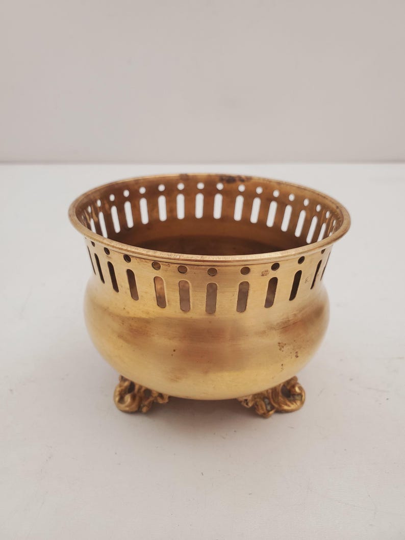 May include: An antique brass bowl with a rounded body and decorative feet. The bowl has a perforated rim and a warm, golden colour. It is likely a decorative piece or a small container for holding items.