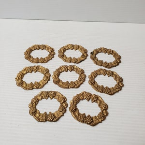May include: Eight antique gold napkin rings. Each ring is oval-shaped and features a detailed design of grapes and leaves, creating a wreath-like appearance. The rings are arranged on a white surface.