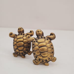 May include: Two gold-coloured turtle figurines with detailed shell patterns. The turtles are facing each other and have their legs outstretched.