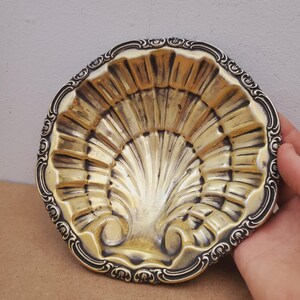 May include: A silver-toned, shell-shaped dish with a decorative, ornate border. The dish has a fan-like pattern in the center, with a slightly raised design. The rim features a detailed, scrolling pattern. The dish is held by a hand.