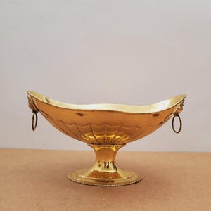May include: A gold-colored decorative bowl with a pedestal base and two ring handles. The bowl has a wide, oval shape with a fluted design on the exterior. The base is round with a detailed, textured edge.