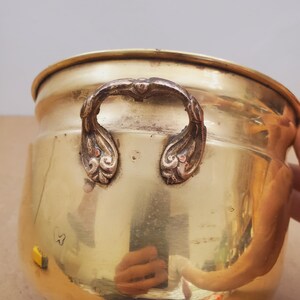 May include: A polished brass planter with ornate handles and a decorative base. The planter has a rounded body and a wide rim. The handles are detailed with floral designs. The brass has a warm, golden hue.