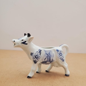 May include: A white ceramic cow creamer with blue floral and windmill designs. The cow has black horns, hooves, and a blue tail. The creamer is a decorative piece for serving milk or cream.