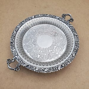May include: A silver-coloured, round serving tray with ornate floral and scrollwork detailing. The tray features two curved handles and a decorative border. The centre of the tray has a detailed floral pattern. The tray is likely made of metal.