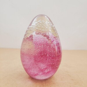 May include: A clear, egg-shaped glass paperweight with a pink, cloud-like design inside. Tiny bubbles are suspended throughout the glass. The paperweight sits on a light brown surface.
