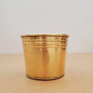 Pode incluir: Um pequeno recipiente cilíndrico de latão com uma base ligeiramente alargada. O recipiente tem faixas horizontais ao redor da parte superior e uma superfície polida e reflexiva. O fundo é de cor neutra.