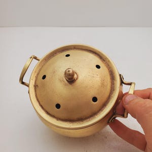 May include: A gold metal incense burner with a lid and two handles. The lid has several small holes for the incense to burn through.