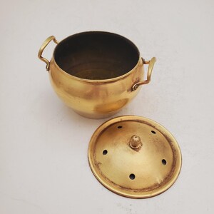 May include: A brass pot with a lid. The pot has two handles and the lid has small holes. The pot and lid are both a shiny gold color.