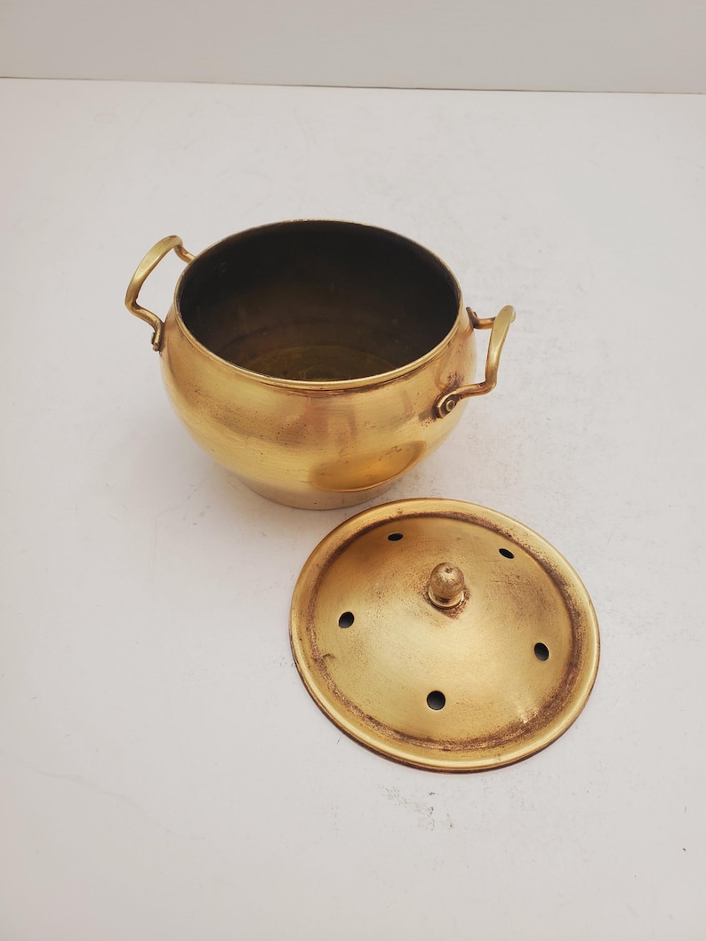 May include: A brass pot with a lid. The pot has two handles and the lid has small holes. The pot and lid are both a shiny gold color.
