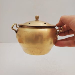 May include: A small, round, brass pot with a lid and two handles. The pot has a smooth, shiny surface and is a light gold color.