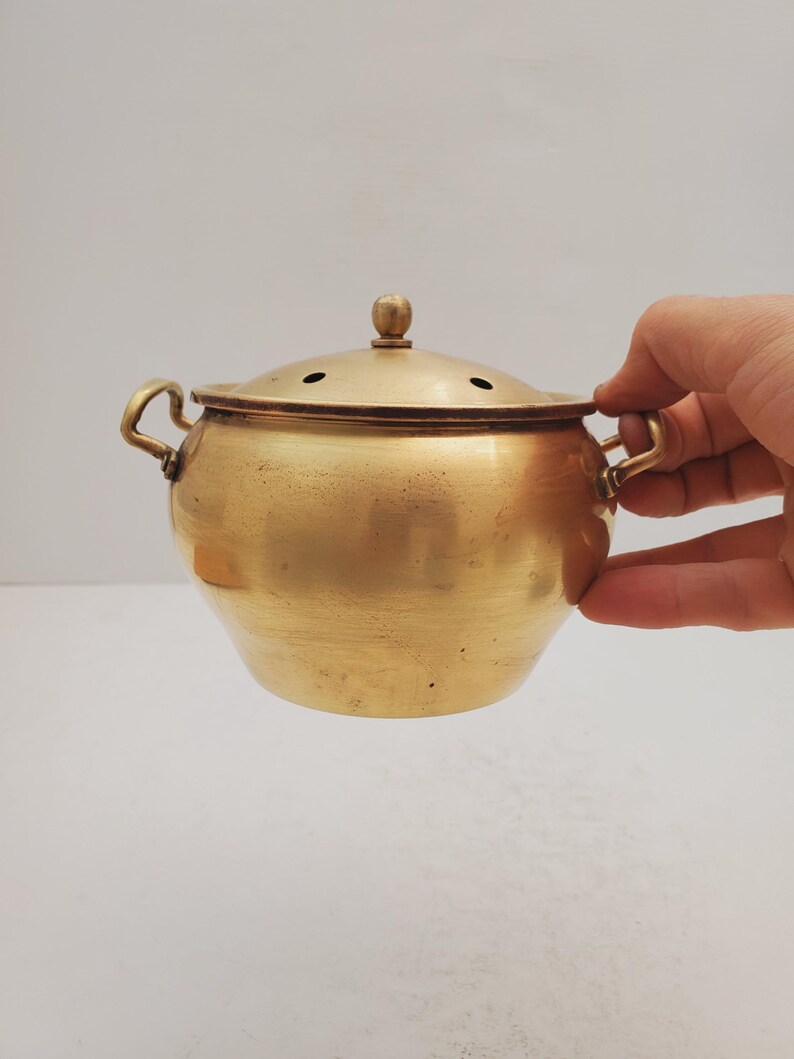 May include: A small, round, brass pot with a lid and two handles. The pot has a smooth, shiny surface and is a light gold color.
