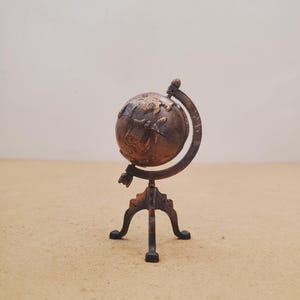May include: A small, metallic, bronze-toned globe model featuring a textured surface with raised continents. The globe is mounted on a curved meridian ring and supported by a three-legged stand, all finished in a matching dark, weathered bronze colour.