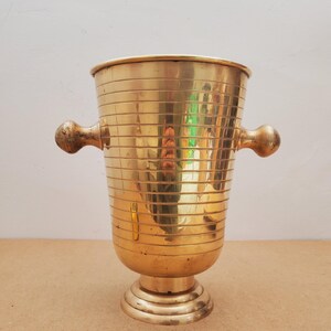 Puede incluir: Un mortero de latón vintage con detalles de ranuras horizontales. El mortero presenta dos asas redondeadas y una base de pedestal. El latón pulido refleja la luz, creando una estética cálida y antigua. Adecuado para la decoración de la cocina.