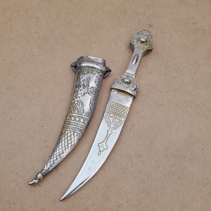 May include: An ornate silver dagger with a decorative sheath. The dagger features intricate carvings on the handle and blade, and the sheath is adorned with detailed patterns. The dagger is displayed on a neutral-toned surface.