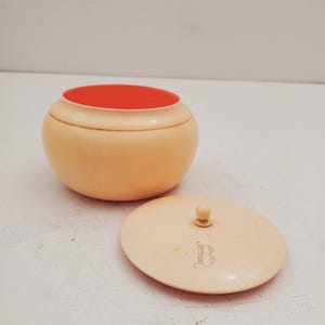 Può includere: Una piccola scatola rotonda di colore crema con un interno rosso e un coperchio con un pomo in cima. Il coperchio ha una lettera 'B' incisa.