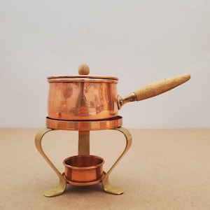 May include: A small copper saucepan with a wooden handle and lid, on a brass stand. The stand has a circular top and a lower ring, supported by curved legs. The pot is designed for heating or serving food.