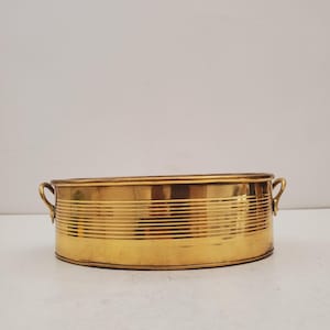 May include: A gold-colored, oval-shaped planter with horizontal etched lines. The planter has two small handles on either side. The planter is made of metal and has a shiny finish. The planter is empty and sits on a white surface.