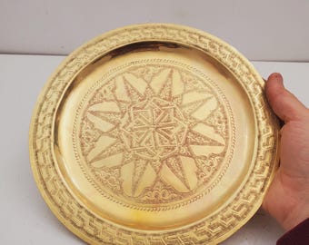 May include: A round, gold-colored metal tray with an intricate starburst design in the center. The tray has a decorative border with a repeating geometric pattern. The tray is held by a person's hand.