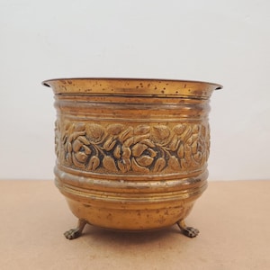 May include: Antique brass planter with floral relief detailing. The planter has a wide rim, a cylindrical body with embossed roses, and rests on three claw feet. The aged brass finish gives it a vintage look.
