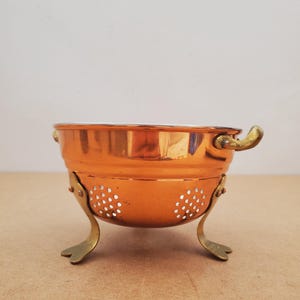 Puede incluir: Un colador de color cobre con asas y patas de latón. El colador tiene un diseño perforado en los laterales. Las asas son curvas y las patas tienen forma de pequeñas hojas. Está diseñado para colocarse sobre una superficie plana.
