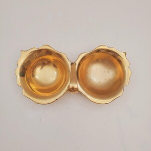 May include: A gold-colored, two-sectioned dish with a connecting handle. Each section is round with a scalloped edge. The dish has a shiny, reflective surface, suggesting it is made of metal.