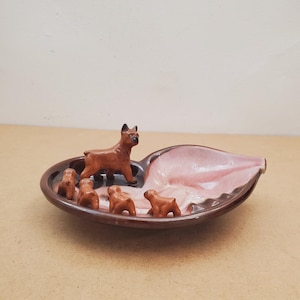 May include: A brown ceramic ashtray in a leaf shape, featuring a brown dog figurine and four smaller dog figurines. The ashtray has a pink interior and a dark brown rim.