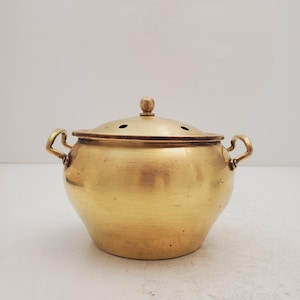 Könnte beinhalten: Ein goldfarbener Topf aus Messing mit Deckel und zwei Griffen. Der Topf hat einen runden Körper und einen leicht gewölbten Deckel mit kleinen Löchern.