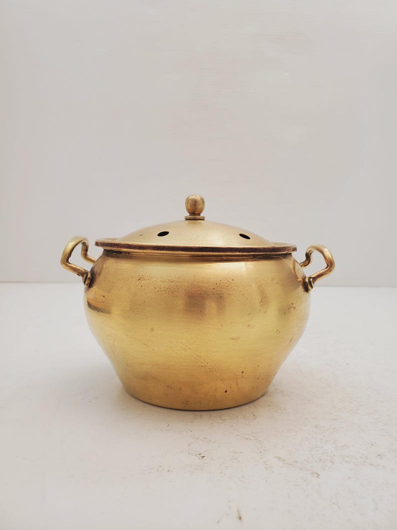 May include: A gold-colored brass pot with a lid and two handles. The pot has a round body and a slightly domed lid with small holes.