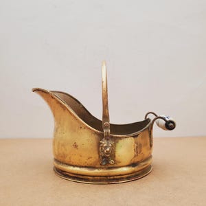 May include: A vintage brass coal scuttle with a handle and a spout. The scuttle has a decorative lion's head on the front and a white and blue ceramic handle. The brass has a warm, aged patina.