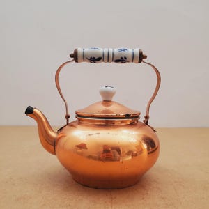 Op de afbeelding: Een koperen theepot met een gebogen tuit en een deksel. De handgreep is gemaakt van koper en heeft een witte keramische greep met blauwe bloemmotieven. De theepot staat op een lichtbruin oppervlak.