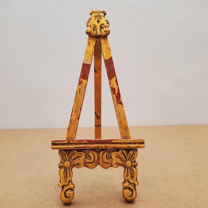 May include: A small, decorative wooden easel with a gold finish and ornate floral carvings. The easel has three legs and a decorative top. The legs have red stripes. The easel is designed to hold a small painting or photograph.