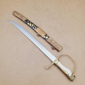 May include: An antique sword with a silver blade and ornate gold and black handle. The hilt is white with gold accents. The scabbard is gold with black and gold detailing. The sword is displayed on a neutral background.