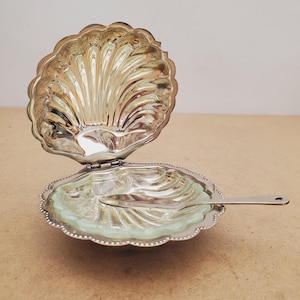 May include: A silver-plated, shell-shaped serving dish with a hinged lid. The dish has a scalloped edge and a light green interior. A small, silver knife rests inside the open dish. The dish is designed to resemble a clam shell.
