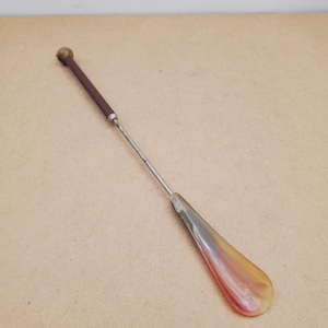 May include: A vintage shoe horn with a brown handle and a metallic shaft. The shoe horn has a curved, amber-colored scoop. The handle has a brass-colored knob at the end.