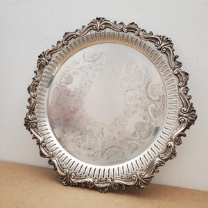 May include: A round silver-coloured serving tray featuring an ornate, scalloped edge with intricate embossed scrollwork and leaf motifs. The central surface displays a detailed engraved pattern of swirling lines, floral elements, and geometric grids.