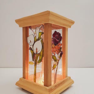 May include: Wooden lantern with stained glass panels depicting white calla lilies and a deep red rose. The lantern has a square base and top, with a warm light shining from inside. The design is reminiscent of a traditional stained glass window.