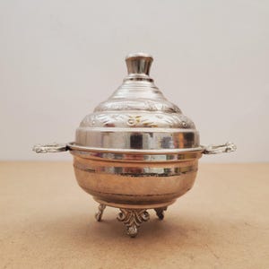 Puede incluir: Un recipiente plateado y ornamentado con tapa, asas pequeñas y patas decorativas. El recipiente tiene una base redondeada y un diseño grabado detallado en la tapa. El diseño general es vintage y elegante.