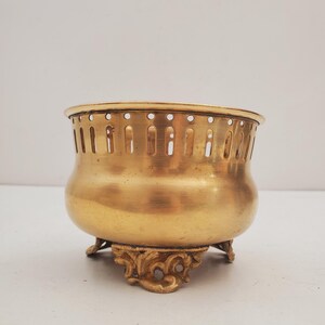 May include: A gold-coloured metal bowl with a rounded body and decorative, ornate feet. The bowl has a series of vertical slots around the top edge. The bowl is likely vintage or antique.