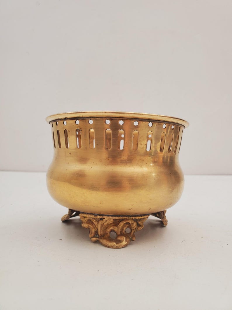 May include: A gold-coloured metal bowl with a rounded body and decorative, ornate feet. The bowl has a series of vertical slots around the top edge. The bowl is likely vintage or antique.