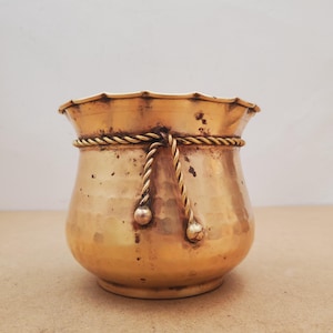 May include: A brass pot with a hammered texture and a scalloped rim. The vessel features a decorative rope-like band encircling the upper section, with two dangling rope ends that terminate in small, rounded spheres on the front surface.